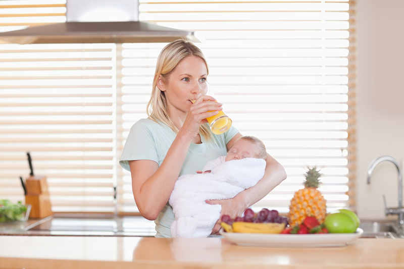 Mlada majka pije zdravi sok i drži bebu koja spava u naručju.